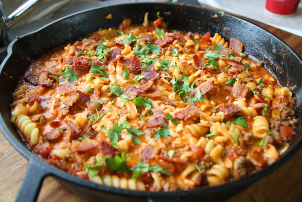 Grandpa's One Pan Ground Beef & Bacon Supper served in a cast iron skillet with rotini pasta, crispy bacon, melted cheddar cheese, and parsley garnish.