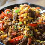 A cast-iron skillet filled with smoky BBQ ground beef, crispy hash browns, bacon, corn, peppers, and melted cheddar cheese with a hearty spoon lift.