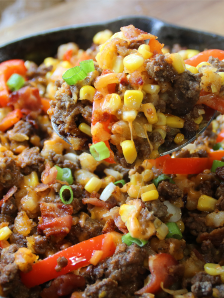 A cast-iron skillet filled with smoky BBQ ground beef, crispy hash browns, bacon, corn, peppers, and melted cheddar cheese with a hearty spoon lift.