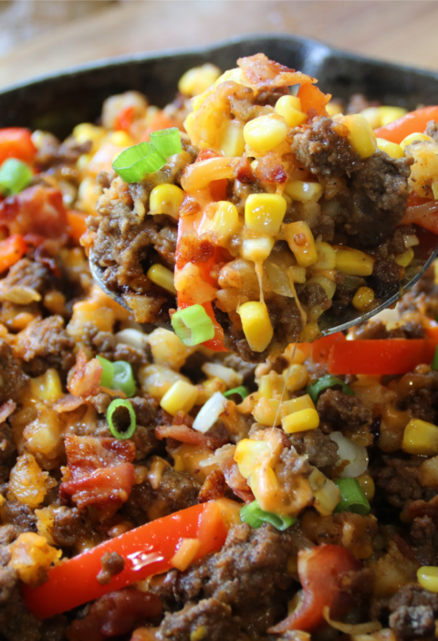 A cast-iron skillet filled with smoky BBQ ground beef, crispy hash browns, bacon, corn, peppers, and melted cheddar cheese with a hearty spoon lift.