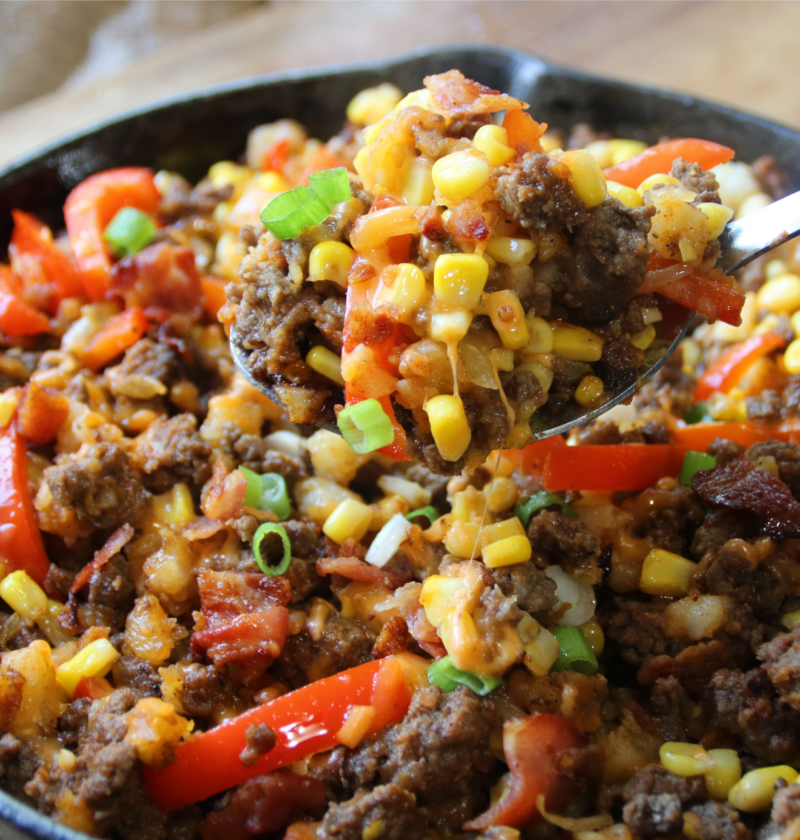 A cast-iron skillet filled with smoky BBQ ground beef, crispy hash browns, bacon, corn, peppers, and melted cheddar cheese with a hearty spoon lift.